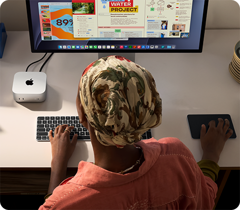 A person typing at a desk with a Mac mini, Studio Display connected, Magic Keyboard, Magic Trackpad, various items