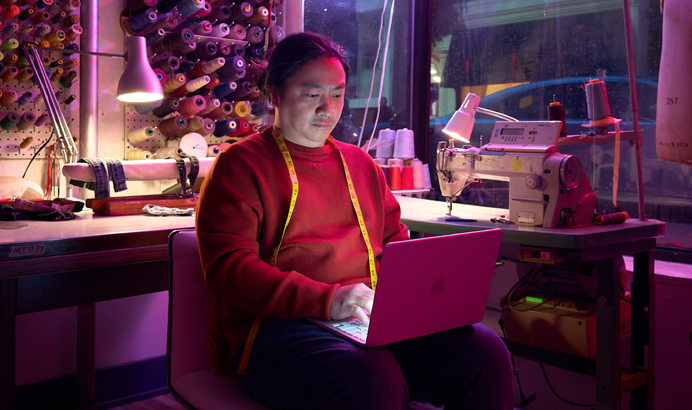 A person uses MacBook Neo, blush color, unplugged on their lap, in a workspace with a sewing machine and related materials, including thread and a dress form