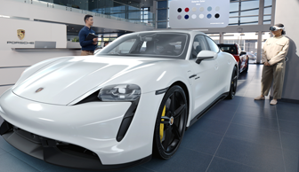 A customer in a Porsche showroom views a realistic 3D projection of a white sportscar with Apple Vision Pro and talks with an employee about color options displayed above the car