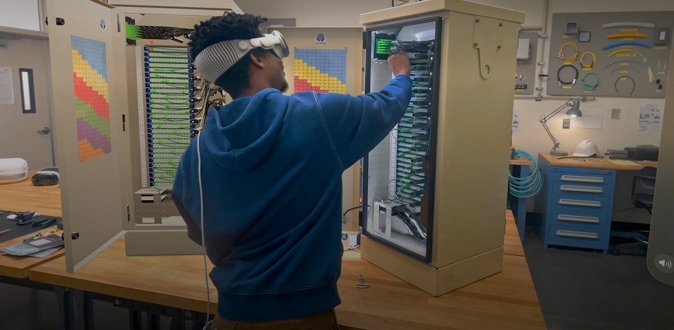 A technician stands at a table and assembles an electrical server tower while wearing Apple Vision Pro and referencing a 3D projection of the tower