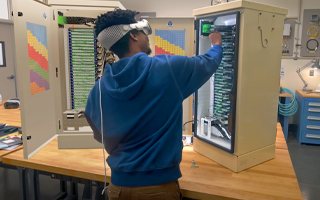 A technician stands at a table and assembles an electrical server tower while wearing Apple Vision Pro and referencing a 3D projection of the tower