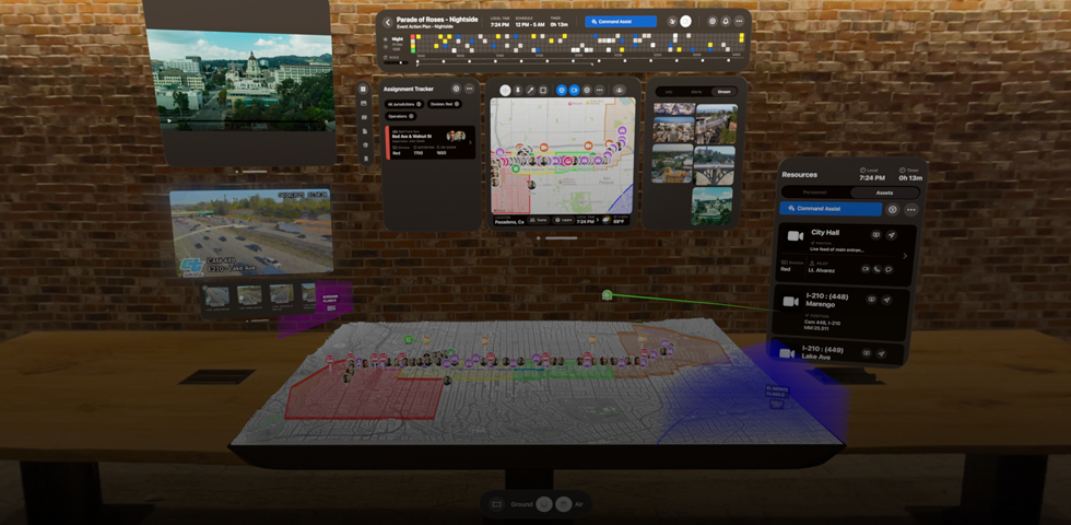 From the view of someone using Apple Vision Pro, a 3D projection of dashboards shows camera feeds, team assignments, and a map of a parade route, in front of a brick wall inside an office