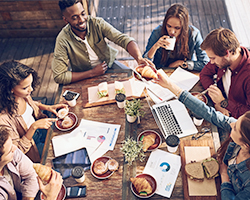 A team of six with laptop and mobile computers collaborate while enjoying a casual lunch.